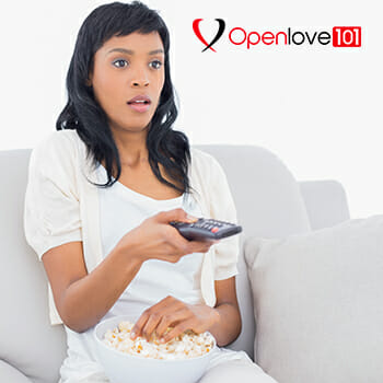 Shocked black haired woman in white clothes watching tv while eating popcorn in a living room